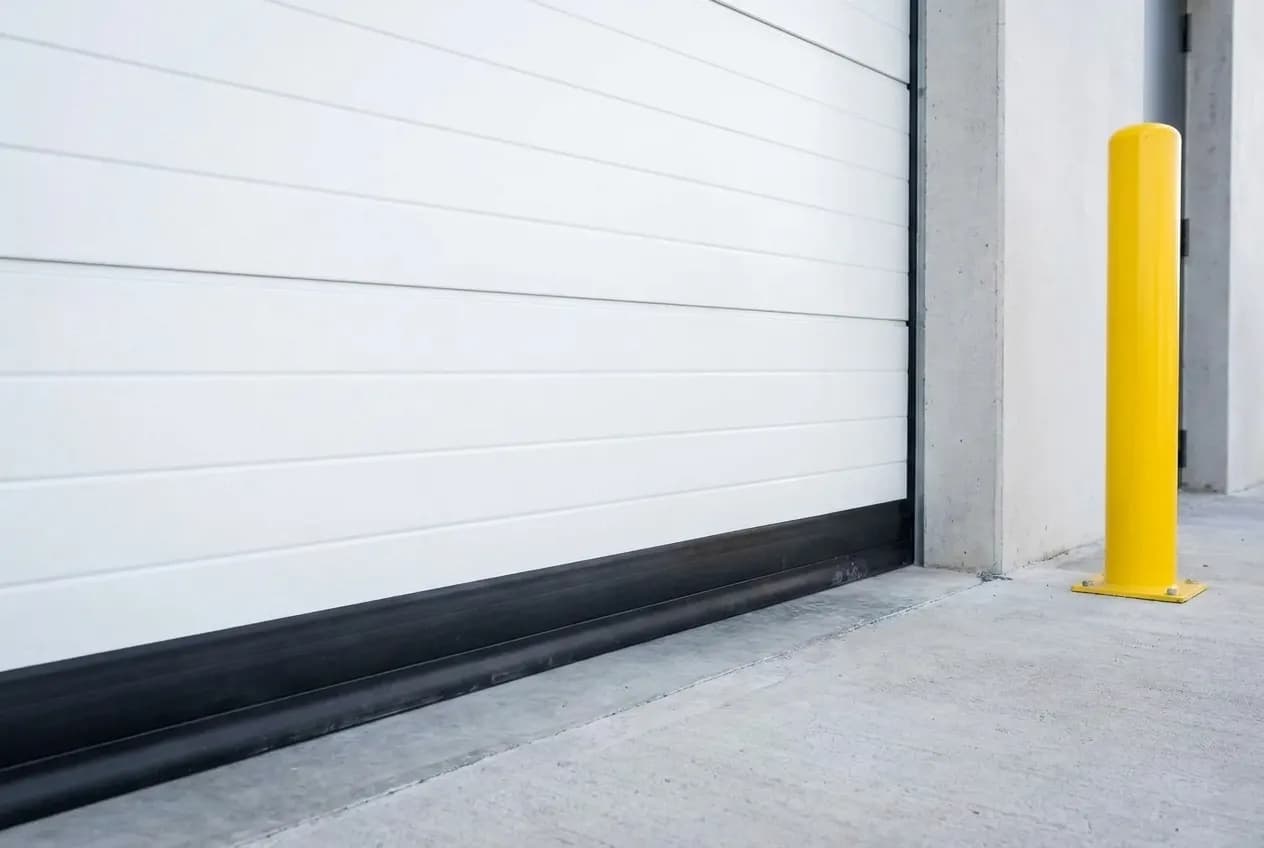 Bas d'une porte sectionnelle industrielle blanche fermée, joint bas noir en caoutchouc posé sur la dalle béton, poteau de protection jaune à côté de la baie
